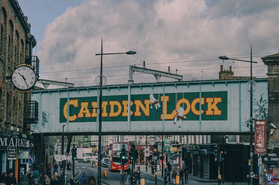 A large, weathered metal bridge with the words 'Camden Lock' painted in bold yellow and red letters spans above a busy street in Camden Market. The bridge is supported by metal beams and has electrical wires running along its length. Below, pedestrians walk across the pavement, some carrying shopping bags, while others stand at street-level shops and stalls. A red double-decker bus is visible in the background, along with various vehicles and street furniture such as traffic lights, streetlamps, and signposts. On the left side, a clock hangs from a brick building that houses a market entrance, with the time showing approximately 11:10. The environment suggests a lively, urban setting during daytime under partly cloudy skies, with natural light illuminating the scene. Camden Town Removals may use this area for furniture transport or home relocation activities, considering the busy street and commercial surroundings.