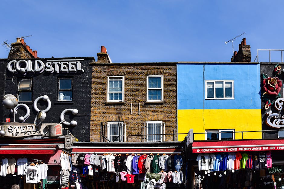 A row of three multi-storey buildings situated behind a street market stall displaying various clothing items, including T-shirts, hoodies, and children’s clothes. The building on the left has a black brick facade with bold white lettering spelling 'COLDSTEEL' on the roof and decorative metallic sculptures attached to the wall. The middle building features a brown brick exterior with three windows, two on the top floor and two on the lower floor, all with white frames. The building on the right is painted in bright blue and yellow, with a window visible on the upper floor and a small opening on the lower level. Above the street market, the sky is clear and blue, and an antenna is visible on the roof of the rightmost building. This scene captures a typical urban setting in Camden with a focus on the exterior walls of the buildings and the merchandise displayed, relevant to house removals, moving logistics, and packing processes that Camden Town Removals may facilitate.