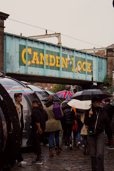A large, weathered metal bridge with the words 'Camden Lock' painted in bold yellow and red letters spans above a busy street in Camden Market. The bridge is supported by metal beams and has electrical wires running along its length. Below, pedestrians walk across the pavement, some carrying shopping bags, while others stand at street-level shops and stalls. A red double-decker bus is visible in the background, along with various vehicles and street furniture such as traffic lights, streetlamps, and signposts. On the left side, a clock hangs from a brick building that houses a market entrance, with the time showing approximately 11:10. The environment suggests a lively, urban setting during daytime under partly cloudy skies, with natural light illuminating the scene. Camden Town Removals may use this area for furniture transport or home relocation activities, considering the busy street and commercial surroundings.